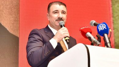 Bir kürsüde konuşma yapan takım elbiseli bir kişi elindeki mikrofonla kalabalığa hitap ediyor. Kürsü üzerinde çeşitli basın kuruluşlarına ait mikrofonlar bulunurken, arka plandaki kırmızı ekran etkinliğe resmi bir atmosfer katıyor. Görsel, bir basın açıklaması veya toplu konuşma anını yansıtıyor.