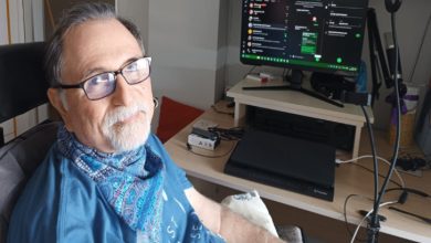 Görselde, bir kişi ev ortamında tıbbi destek ekipmanlarıyla birlikte bilgisayar başında oturuyor. Çalışma masasında ekran, teknik cihazlar ve kayıt ekipmanına benzer araçlar yer alırken; sahne, günlük yaşamın üretim, iletişim ve teknolojik destekle sürdürüldüğü bir anı yansıtıyor.