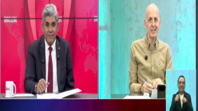 Bir televizyon programında iki sunucu veya konuşmacı, farklı stüdyo ekranlarından canlı yayında sohbet ediyor. Görüntünün sağ alt köşesinde işaret dili tercümanı yer alırken, arka planda kırmızı ve turkuaz tonlarında stüdyo dekoru bulunuyor. Katılımcılar masalarında not alırken ekrana bakıyor.