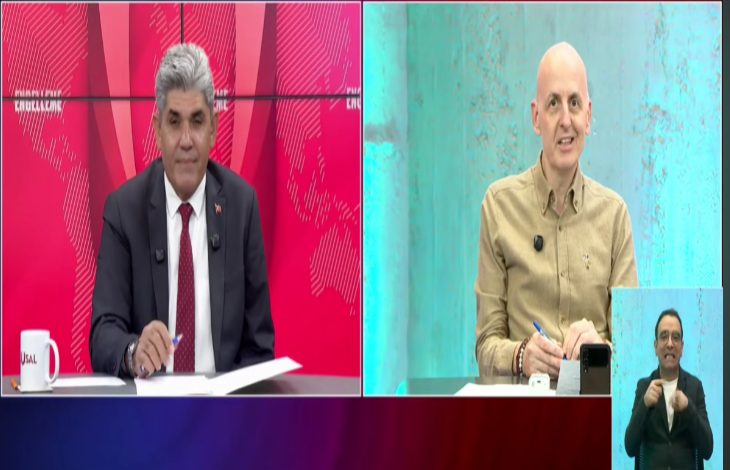 Bir televizyon programında iki sunucu veya konuşmacı, farklı stüdyo ekranlarından canlı yayında sohbet ediyor. Görüntünün sağ alt köşesinde işaret dili tercümanı yer alırken, arka planda kırmızı ve turkuaz tonlarında stüdyo dekoru bulunuyor. Katılımcılar masalarında not alırken ekrana bakıyor.