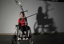 Görselde, tekerlekli sandalye kullanan genç bir sporcu, elinde yay ve oklarla okçuluk antrenmanı yaparken görülüyor. Karanlık fonda çekilen fotoğrafta sporcunun duvara yansıyan gölgesi dikkat çekiyor. Görsel, okçuluk sporuyla ilgilenen sporcunun antrenman sürecini ve odaklanmış duruşunu yansıtıyor.
