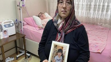 Bir ev ortamında çekilen fotoğrafta, başörtülü bir kadın elinde çerçeveli bir çocuk fotoğrafı tutarak kameraya bakıyor. Arka planda ise tıbbi cihazların bulunduğu bir yatakta yatan bir çocuk görülüyor. Görsel, sağlık sorunlarıyla mücadele eden bir çocuğun ve ailesinin yaşadığı zorlu sürece dikkat çekiyor.