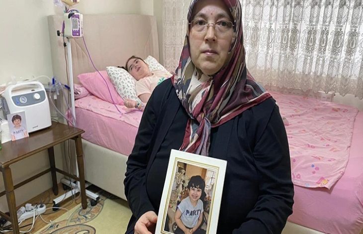 Bir ev ortamında çekilen fotoğrafta, başörtülü bir kadın elinde çerçeveli bir çocuk fotoğrafı tutarak kameraya bakıyor. Arka planda ise tıbbi cihazların bulunduğu bir yatakta yatan bir çocuk görülüyor. Görsel, sağlık sorunlarıyla mücadele eden bir çocuğun ve ailesinin yaşadığı zorlu sürece dikkat çekiyor.