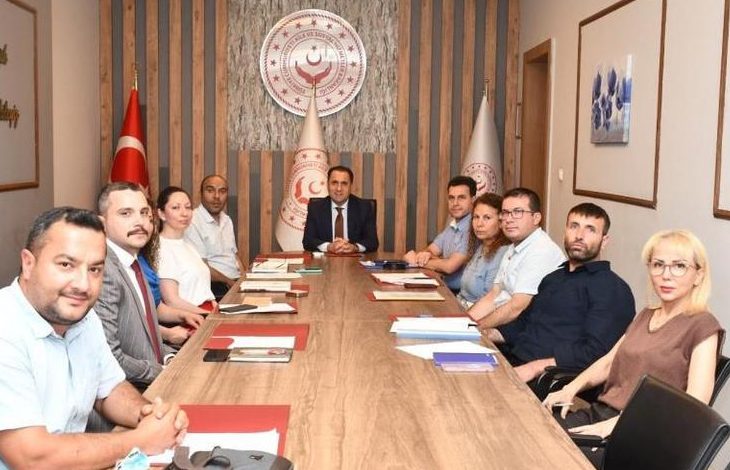 Bir toplantı salonunda uzun bir masa etrafında oturan dokuz kişi kameraya bakarak poz veriyor. Masanın başında bir yönetici yer alırken, arka duvarda Bakanlık logosu ve Türk bayrağı bulunuyor. Görsel, resmi bir değerlendirme veya koordinasyon toplantısını yansıtıyor.
