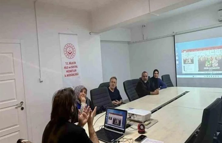 Görselde, bir toplantı salonunda gerçekleştirilen eğitim programı görülüyor. Masanın etrafında oturan katılımcılar, projeksiyon cihazıyla yansıtılan sunumu takip ediyor. Salonda bulunan afişte, programın T.C. Bilecik Aile ve Sosyal Hizmetler İl Müdürlüğü tarafından düzenlendiği anlaşılıyor. Eğitim ortamında bilgilendirme ve farkındalık çalışması yapıldığı görülüyor.