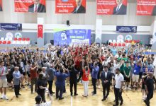 Bu görselde, bir spor salonunda düzenlenen kalabalık bir etkinlik sırasında katılımcılar ellerini havaya kaldırarak coşkulu bir şekilde kutlama yaparken, salonun ortasında bulunan görevliler ve sporcular etkinliğe eşlik etmektedir.