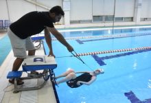 Fotoğrafta bir yüzme havuzunda antrenman anı görülüyor. Havuz kenarında duran bir antrenör, su içindeki sporcuya yönlendirme yapıyor. Sporcu sırtüstü pozisyonda yüzeye yakın şekilde dururken, sahne performans sporu ile bireysel gelişim ve destekleyici antrenman sürecini yansıtıyor.