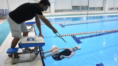 Fotoğrafta bir yüzme havuzunda antrenman anı görülüyor. Havuz kenarında duran bir antrenör, su içindeki sporcuya yönlendirme yapıyor. Sporcu sırtüstü pozisyonda yüzeye yakın şekilde dururken, sahne performans sporu ile bireysel gelişim ve destekleyici antrenman sürecini yansıtıyor.