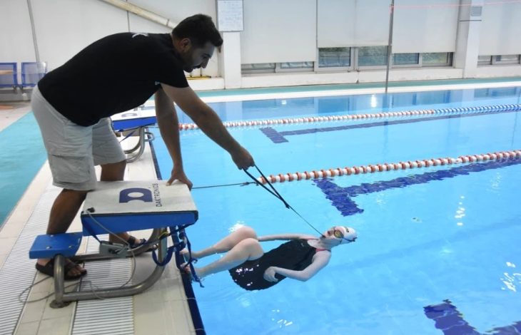 Fotoğrafta bir yüzme havuzunda antrenman anı görülüyor. Havuz kenarında duran bir antrenör, su içindeki sporcuya yönlendirme yapıyor. Sporcu sırtüstü pozisyonda yüzeye yakın şekilde dururken, sahne performans sporu ile bireysel gelişim ve destekleyici antrenman sürecini yansıtıyor.