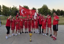 Görselde, kırmızı milli takım formaları giyen sporcular ve teknik ekip, ellerindeki madalyalarla Türk bayrağı önünde toplu fotoğraf veriyor. Ön planda kupa yer alırken, takımın şampiyona sonrası elde ettiği dereceyi birlikte kutladığı gururlu ve coşkulu bir an yansıtılıyor.