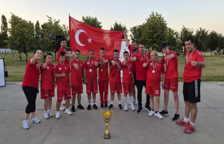 Görselde, kırmızı milli takım formaları giyen sporcular ve teknik ekip, ellerindeki madalyalarla Türk bayrağı önünde toplu fotoğraf veriyor. Ön planda kupa yer alırken, takımın şampiyona sonrası elde ettiği dereceyi birlikte kutladığı gururlu ve coşkulu bir an yansıtılıyor.