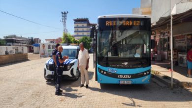 Trafik polisi yol kenarında duran mavi renkli bir şehir otobüsünün önünde şoförle konuşurken, yanında park etmiş bir polis aracı ve çevrede dükkânların bulunduğu güneşli bir cadde görülmektedir.