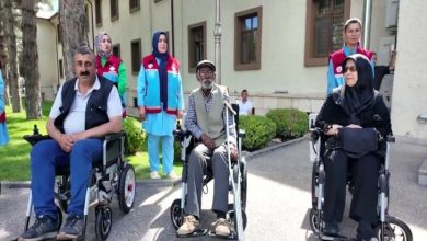Görselde, bir bina önünde düzenlenen teslim töreninde akülü tekerlekli sandalye kullanan üç engelli birey yan yana oturuyor. Arkalarında görevli oldukları anlaşılan üniformalı dört kişi ayakta dururken, çevrede yeşil alan ve bina girişi görülüyor. Fotoğraf, ihtiyaç sahibi engelli bireylere akülü tekerlekli sandalye desteği sağlanan bir etkinlikten alınmış izlenimi veriyor.