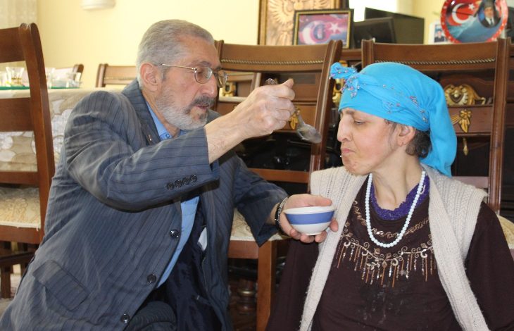 Ev ortamında çekilen fotoğrafta, yaşlı bir erkek bir kâşıkla yanındaki kadına yemek yediriyor. Mavi başörtüsü takan kadın sandalyede otururken, erkek elindeki kâseyle ona destek oluyor. Görsel, bakım, fedakârlık ve aile içi dayanışmayı yansıtan duygusal bir anı gösteriyor.