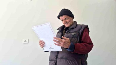 Görselde, bere ve şişme yelek giyen bir kişi elindeki belgeyi dikkatle incelerken görülüyor. Sade ve açık renkli bir odada çekilen fotoğrafta kişinin yüzünde memnuniyet ve rahatlama ifadesi dikkat çekiyor. Belgeye odaklanan kişi ayakta dururken, görüntü önemli bir gelişme veya sevindirici bir haber aldığı anı yansıtıyor.