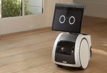 Ahşap zeminde duran, ekranında iki büyük dairesel göz bulunan sevimli görünümlü küçük bir ev robotu, geniş bir pencerenin önünde bekliyor.