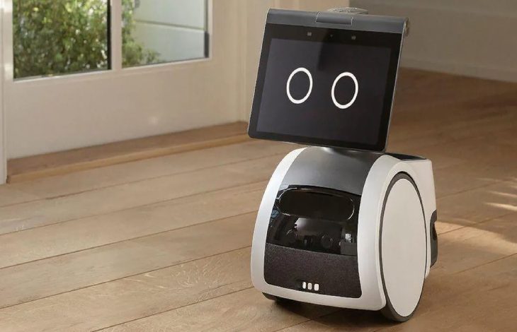 Ahşap zeminde duran, ekranında iki büyük dairesel göz bulunan sevimli görünümlü küçük bir ev robotu, geniş bir pencerenin önünde bekliyor.