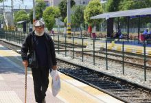 Elinde baston ve alışveriş poşeti taşıyan Ali Bektaş, açık havadaki bir tren istasyonunda rayların yanında yürümektedir.