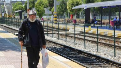 Elinde baston ve alışveriş poşeti taşıyan Ali Bektaş, açık havadaki bir tren istasyonunda rayların yanında yürümektedir.