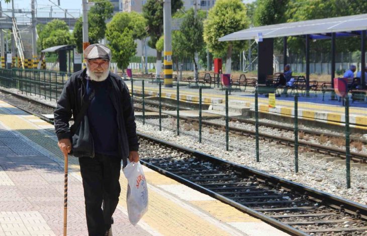 Elinde baston ve alışveriş poşeti taşıyan Ali Bektaş, açık havadaki bir tren istasyonunda rayların yanında yürümektedir.