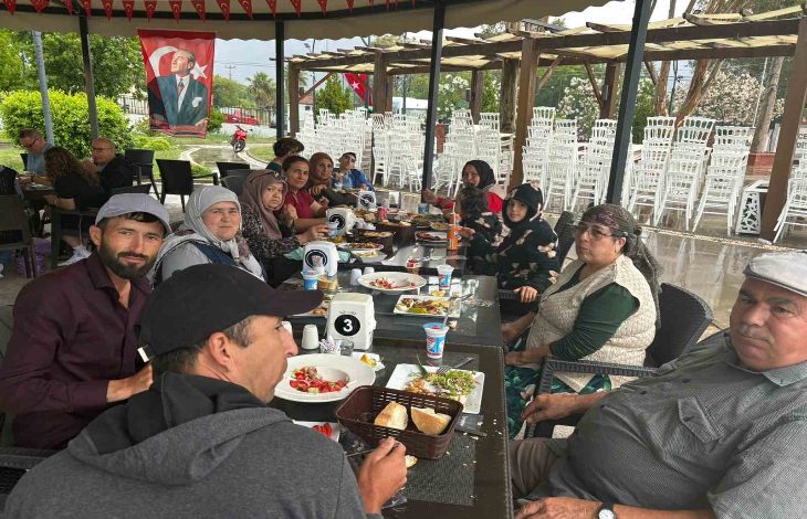 Görselde, açık alanda düzenlenen bir etkinlikte uzun bir masa etrafında bir araya gelen aileler yemek eşliğinde vakit geçiriyor. Türk bayrakları ve doğayla iç içe ortam dikkat çekerken, katılımcıların birlikte zaman geçirdiği samimi ve sosyal bir buluşma atmosferi öne çıkıyor.
