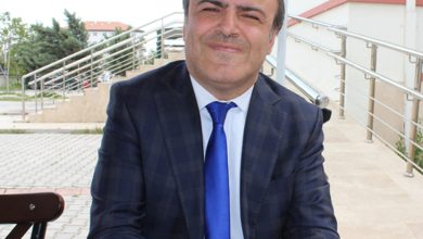 Takım elbiseli bir kişi, dış mekânda merdiven kenarında oturuyor; arka planda açık renk bina ve yeşillik görünüyor.