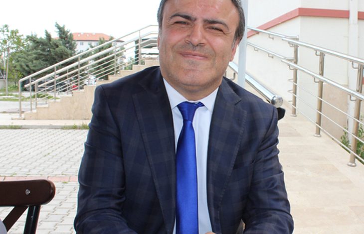Takım elbiseli bir kişi, dış mekânda merdiven kenarında oturuyor; arka planda açık renk bina ve yeşillik görünüyor.