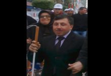 Görüntüde takım elbiseli bir kişi, elinde beyaz bastonla kalabalık bir ortamda yürürken görülüyor. Çevresinde yer alan kişilerle birlikte bir etkinlik veya yürüyüşe katıldığı izlenimi veren kare, açık havada çekilmiş.