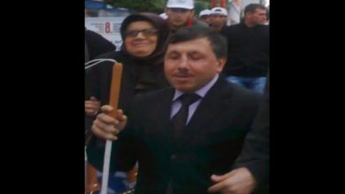 Görüntüde takım elbiseli bir kişi, elinde beyaz bastonla kalabalık bir ortamda yürürken görülüyor. Çevresinde yer alan kişilerle birlikte bir etkinlik veya yürüyüşe katıldığı izlenimi veren kare, açık havada çekilmiş.