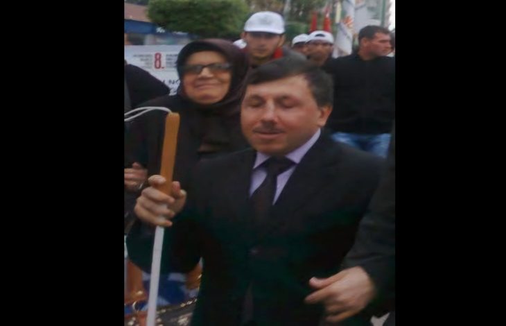 Görüntüde takım elbiseli bir kişi, elinde beyaz bastonla kalabalık bir ortamda yürürken görülüyor. Çevresinde yer alan kişilerle birlikte bir etkinlik veya yürüyüşe katıldığı izlenimi veren kare, açık havada çekilmiş.