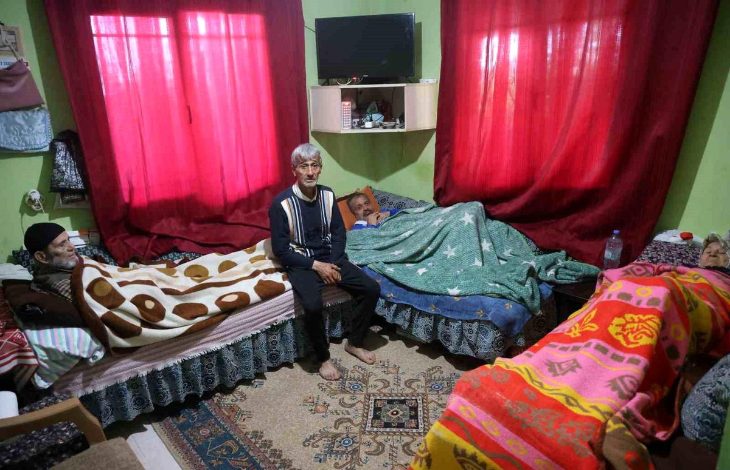 Görselde, mütevazı bir ev ortamında aynı odada kalan yaşlı bireyler ve ortada oturan bir kişi görülüyor. Yatakların yan yana yer aldığı odada bakım ve birlikte yaşam düzeni dikkat çekerken, sahne aile içi dayanışma, sorumluluk ve uzun süreli bakımın günlük yaşamını yansıtıyor.