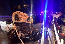 Gece saatlerinde meydana gelen trafik kazasının ardından çekilen fotoğrafta, ön kısmı ağır hasar görmüş beyaz bir otomobil yol üzerinde duruyor. Aracın kaputu tamamen açılmış ve motor bölümü büyük ölçüde zarar görmüş durumda. Yolun sağ tarafında bariyerlerin üzerine çıkmış başka bir otomobil görülürken, olay yerinde polis ve acil müdahale ekiplerinin araçlarından yansıyan kırmızı ve mavi ışıklar dikkat çekiyor. Islak yol yüzeyi ve çevredeki görevliler, kazanın ardından yürütülen çalışmaları yansıtıyor.