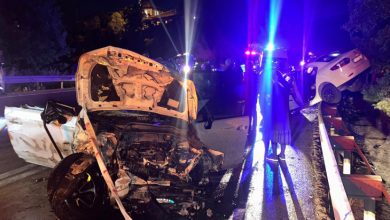 Gece saatlerinde meydana gelen trafik kazasının ardından çekilen fotoğrafta, ön kısmı ağır hasar görmüş beyaz bir otomobil yol üzerinde duruyor. Aracın kaputu tamamen açılmış ve motor bölümü büyük ölçüde zarar görmüş durumda. Yolun sağ tarafında bariyerlerin üzerine çıkmış başka bir otomobil görülürken, olay yerinde polis ve acil müdahale ekiplerinin araçlarından yansıyan kırmızı ve mavi ışıklar dikkat çekiyor. Islak yol yüzeyi ve çevredeki görevliler, kazanın ardından yürütülen çalışmaları yansıtıyor.