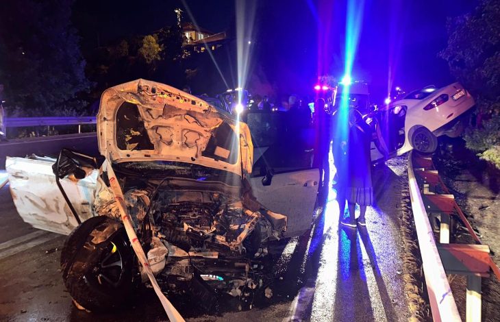 Gece saatlerinde meydana gelen trafik kazasının ardından çekilen fotoğrafta, ön kısmı ağır hasar görmüş beyaz bir otomobil yol üzerinde duruyor. Aracın kaputu tamamen açılmış ve motor bölümü büyük ölçüde zarar görmüş durumda. Yolun sağ tarafında bariyerlerin üzerine çıkmış başka bir otomobil görülürken, olay yerinde polis ve acil müdahale ekiplerinin araçlarından yansıyan kırmızı ve mavi ışıklar dikkat çekiyor. Islak yol yüzeyi ve çevredeki görevliler, kazanın ardından yürütülen çalışmaları yansıtıyor.