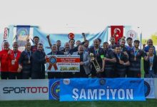 Şampiyonluk kupasını ve ödül çekini birlikte kaldıran madalyalı futbolcular ile teknik ekip, 'Şampiyon' yazılı pankart önünde sevinçle poz vererek elde ettikleri başarıyı kutlamakta.