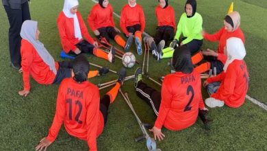Fotoğrafta ampute kadın futbolcuların sahada daire şeklinde oturarak antrenman veya takım buluşması yaptığı görülüyor. Sporcular turuncu formalar giyiyor ve ortada bir futbol topu bulunuyor. Bazı sporcular protez veya destek ekipmanlarıyla yer alırken, görüntü takım ruhu, dayanışma ve spora katılım temasını öne çıkarıyor.