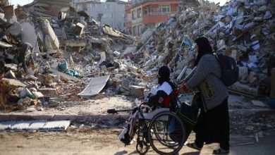 Görselde, tekerlekli sandalyedeki bir kişi ve ona eşlik eden bir kadın, yıkılmış binaların enkazları arasında ilerliyor. Fotoğraf, afetlerin engelli bireyler üzerindeki etkisini ve erişilebilir afet yönetiminin önemini vurgulayan çarpıcı bir sahne sunuyor.