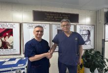 Fotoğrafta iki sağlık çalışanı veya doktor, hastane ortamında el sıkışırken görülüyor. Arkalarında tıbbi eğitim ve kardiyoloji alanıyla ilgili posterler yer alıyor. Görsel, uluslararası mesleki iş birliği, eğitim veya bilgi paylaşımını temsil eden resmi bir ziyaret anını yansıtıyor.