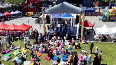 Açık havada düzenlenen etkinlikte insanlar renkli minderlerin üzerinde oturarak sahnedeki programı izlerken, çevrede kurulu çadırlar ve kalabalık bir festival atmosferi dikkat çekmektedir.