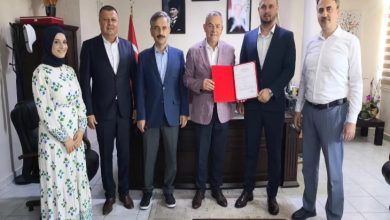 Bir ofis ortamında altı kişi birlikte poz veriyor. Ortadaki iki kişi kırmızı dosya içinde bir belge tutuyor; arka planda Türk bayrağı ve duvarda çerçeveli portreler görülüyor. Görüntü resmi bir belge teslimi ya da açılış/izin töreni izlenimi veriyor.