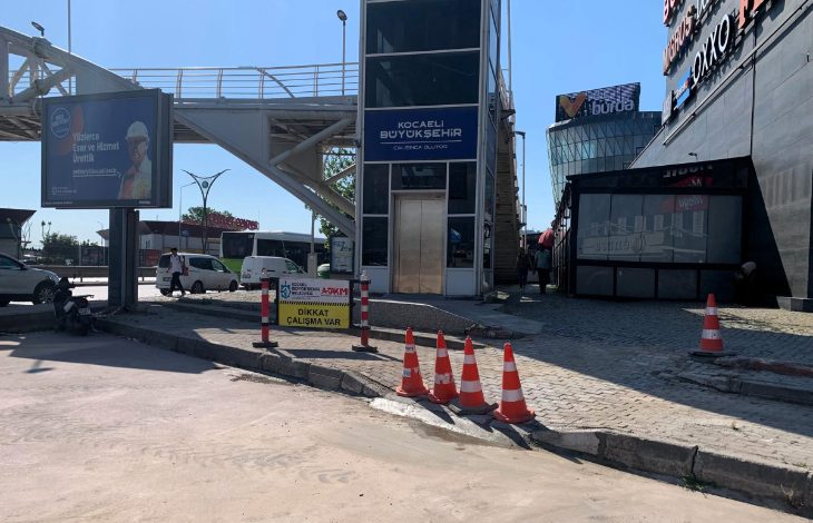 Yaya üst geçidinin yanında bulunan asansör kulesi önünde sürdürülen kaldırım ve yol çalışması görülüyor. Çalışma alanı trafik konileri ve uyarı levhalarıyla çevrilirken, bölgede erişilebilirlik düzenlemelerine yönelik çalışmalar dikkat çekiyor.