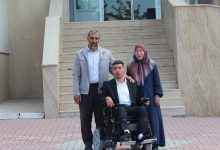 Fotoğrafta bir binanın girişindeki merdivenlerin önünde üç kişi poz veriyor. Ortada, motorlu tekerlekli sandalyede oturan genç bir erkek bulunuyor. Yanında ayakta duran bir kadın ve bir erkek, gencin omzuna destekleyici şekilde dokunuyor. Genç erkek takım elbise giyerken, yanındaki kişiler günlük kıyafetler içinde görünüyor. Arka planda geniş merdivenler ve cam kapılı bir bina girişi yer alıyor. Görsel, mezuniyet veya önemli bir başarı sonrası çekilmiş bir aile hatırası fotoğrafı izlenimi veriyor.