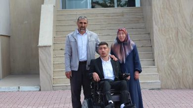 Fotoğrafta bir binanın girişindeki merdivenlerin önünde üç kişi poz veriyor. Ortada, motorlu tekerlekli sandalyede oturan genç bir erkek bulunuyor. Yanında ayakta duran bir kadın ve bir erkek, gencin omzuna destekleyici şekilde dokunuyor. Genç erkek takım elbise giyerken, yanındaki kişiler günlük kıyafetler içinde görünüyor. Arka planda geniş merdivenler ve cam kapılı bir bina girişi yer alıyor. Görsel, mezuniyet veya önemli bir başarı sonrası çekilmiş bir aile hatırası fotoğrafı izlenimi veriyor.