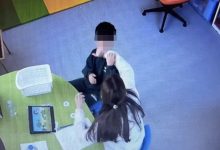 Güvenlik kamerası açısından çekilen görselde bir çocuk ve yanında oturan bir yetişkin görülüyor. İç mekânda masa başında gerçekleşen bir etkileşim anı kaydedilmiş; masanın üzerinde tablet ve çeşitli materyaller bulunuyor. Görüntü, eğitim veya bakım ortamını andıran bir alanı gösteriyor.