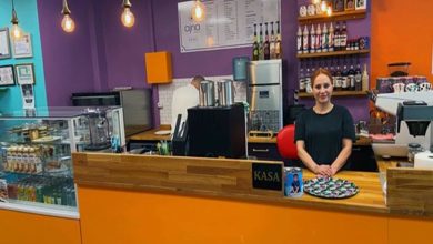 Renkli bir kafe ortamında tezgâhın arkasında duran bir kişi kameraya bakıyor. Mor ve turuncu tonların hâkim olduğu mekânda kahve ekipmanları, içecek şişeleri ve solda cam vitrin içinde ürünler yer alıyor. Genel görünüm küçük ve canlı bir kafe atmosferi oluşturuyor.