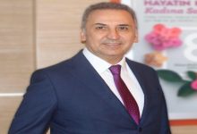 Görselde takım elbiseli bir kişi iç mekânda ayakta dururken görülüyor. Lacivert takım elbise, beyaz gömlek ve bordo desenli kravat giyen kişi kameraya doğru hafifçe gülümseyerek poz veriyor. Arka planda pembe çiçek görsellerinin yer aldığı ve kadın sağlığına ilişkin olduğu anlaşılan bir afiş bulunuyor. Fotoğraf profesyonel bir portre çekimi izlenimi veriyor.