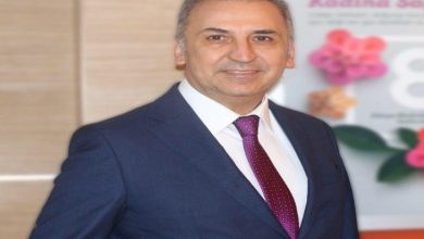 Görselde takım elbiseli bir kişi iç mekânda ayakta dururken görülüyor. Lacivert takım elbise, beyaz gömlek ve bordo desenli kravat giyen kişi kameraya doğru hafifçe gülümseyerek poz veriyor. Arka planda pembe çiçek görsellerinin yer aldığı ve kadın sağlığına ilişkin olduğu anlaşılan bir afiş bulunuyor. Fotoğraf profesyonel bir portre çekimi izlenimi veriyor.