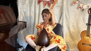 Nil Korkmaz sahnede sandalyede oturuyor, elinde bağlama tutuyor; yanında piyano ve gitar var.