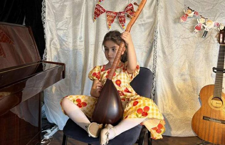 Nil Korkmaz sahnede sandalyede oturuyor, elinde bağlama tutuyor; yanında piyano ve gitar var.