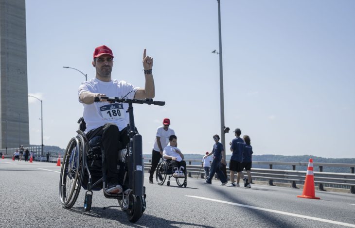 Yarış numarası göğsünde bulunan tekerlekli sandalye kullanan bir kişi önüne monte edilmiş elektrikli bir aparatla ilerlerken elini kaldırıp işaret veriyor; arka planda ise diğer katılımcılar ve görevliler görülüyor.